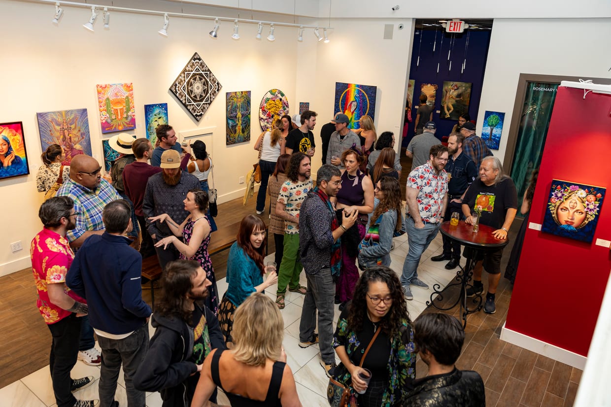 A large crowd of adults mingles at an art gallery opening, surrounded by colorful paintings hung on white and red walls.
