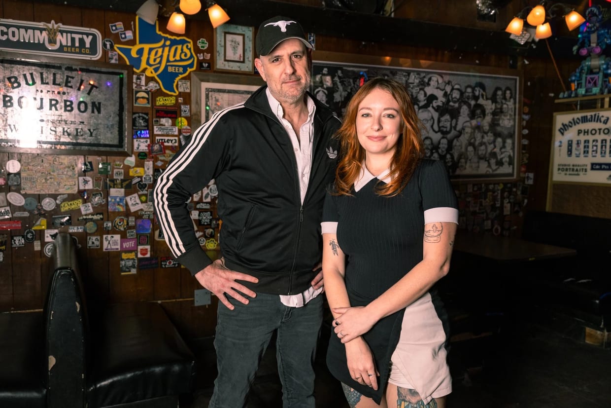 A man and a woman stand together inside a dimly lit bar decorated with beer signs, stickers, and memorabilia.