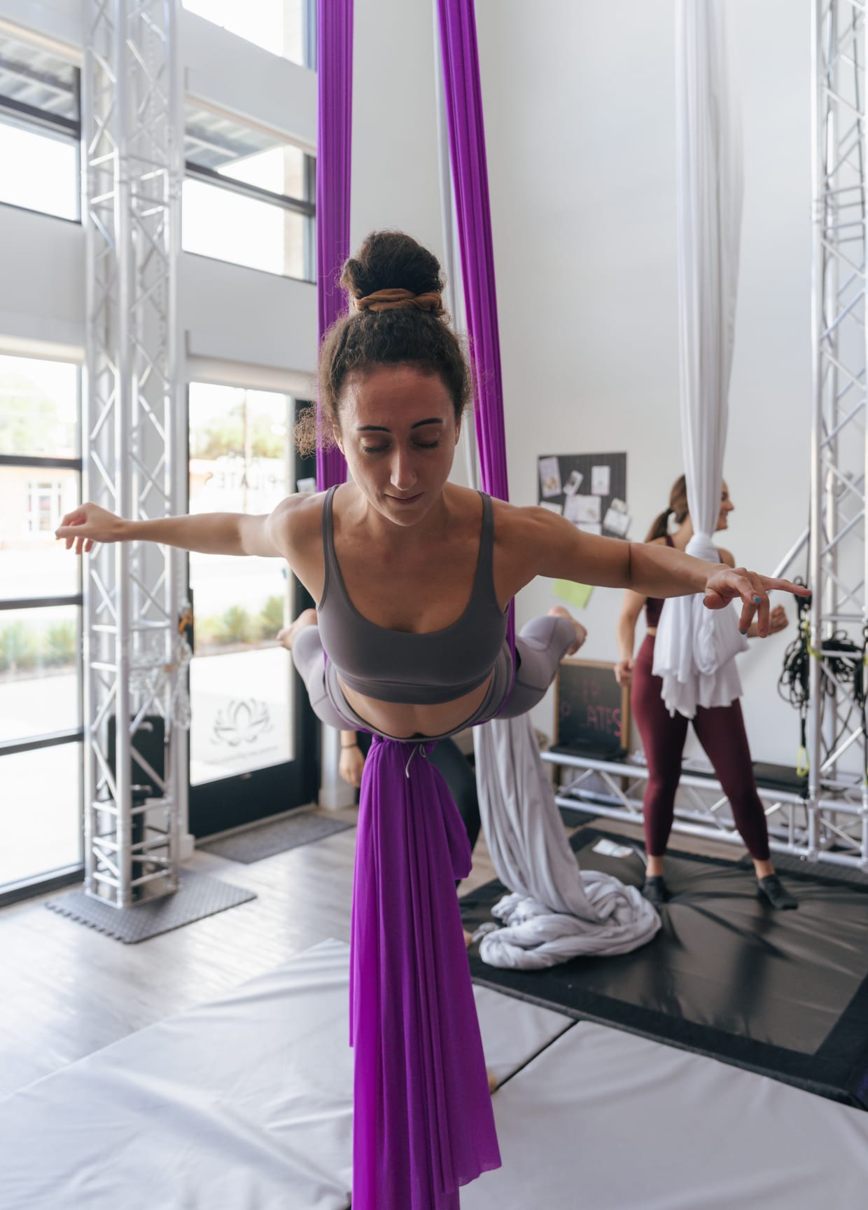 A woman holds an aerial silks pose above a mat in a bright studio while another person works with white silks behind her.