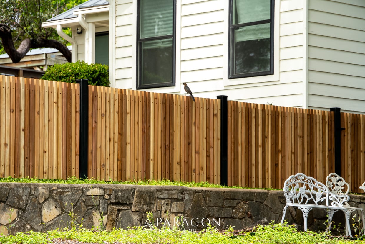 A newly installed cedar privacy fence with black metal posts runs along a stone retaining wall beside a two-story white house, with a bird perched on top and a white iron garden bench visible at lower right.