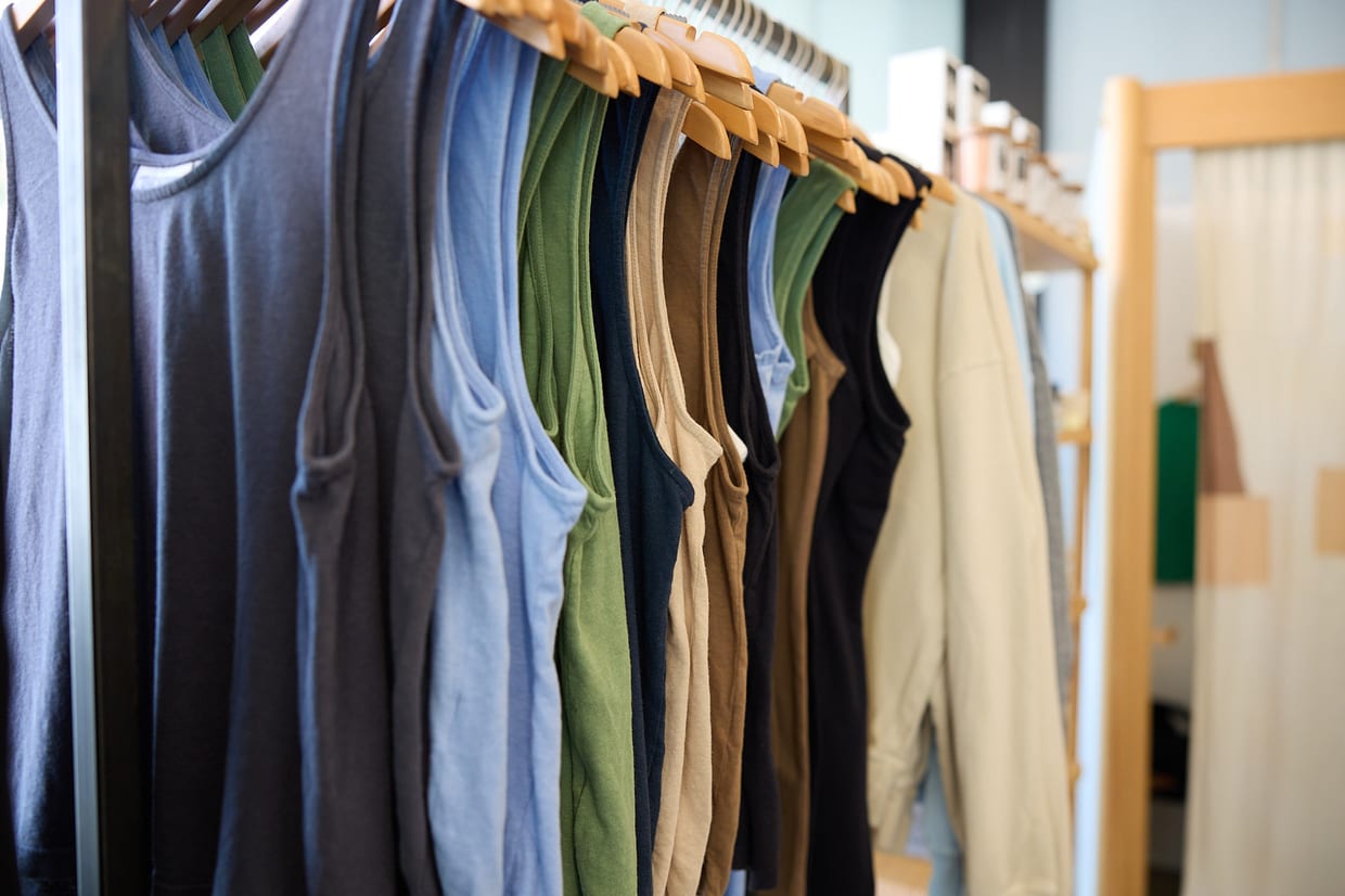 A row of sleeveless tank tops in various earth and neutral colors hanging on wooden hangers on a clothing rack inside a retail shop.