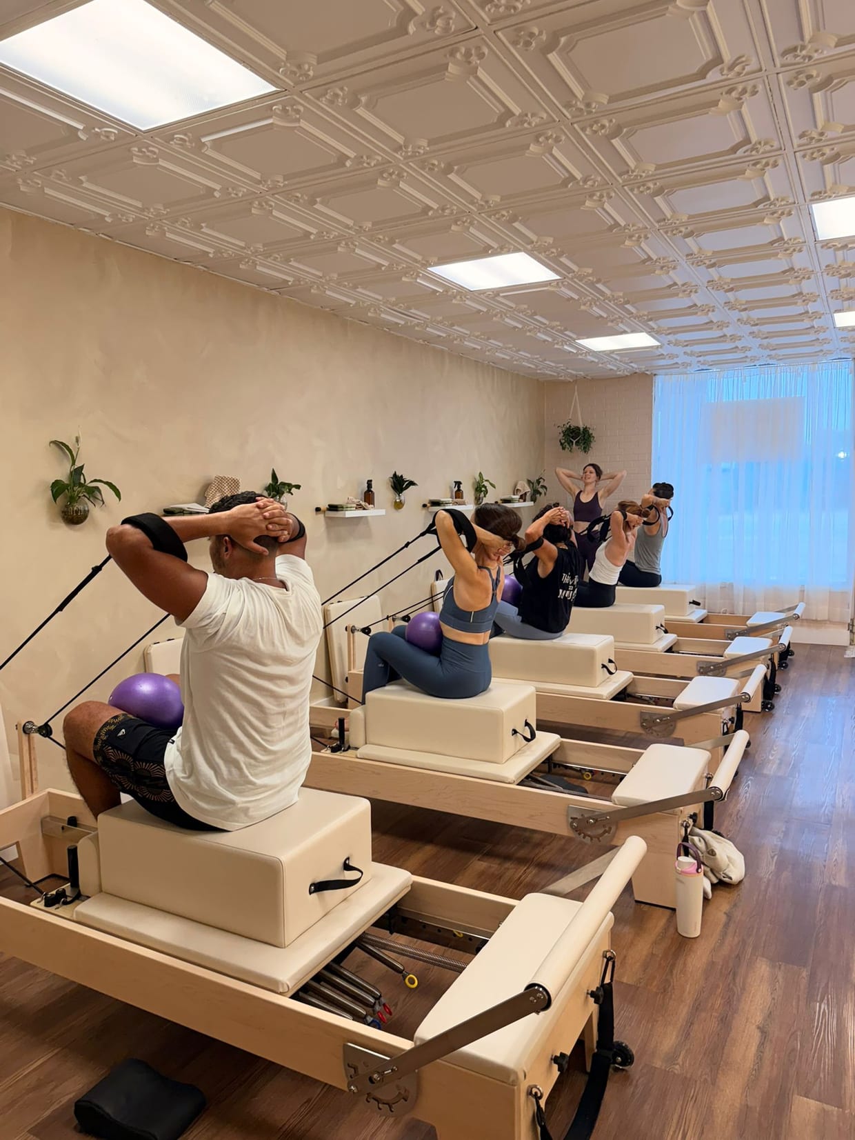 A group of clients perform a seated abdominal exercise on Pilates reformers in a bright studio with an ornate tin ceiling and wood floors.