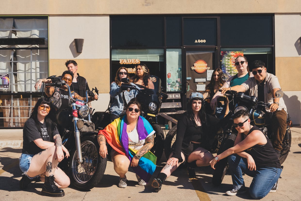 The Reunion Barbershop team gathered outside the shop with their motorcycles, one of them wrapped in a Pride flag, under the window signs and the shop’s logo on the door.