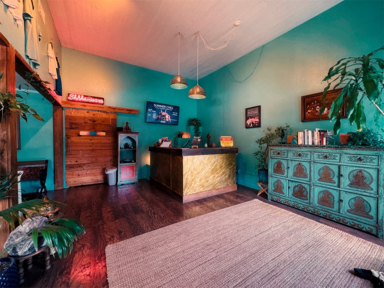 A yoga studio reception area with teal walls, a gold-paneled front desk, ornate turquoise cabinet, pendant lights, houseplants, and a flat-screen display mounted on the wall