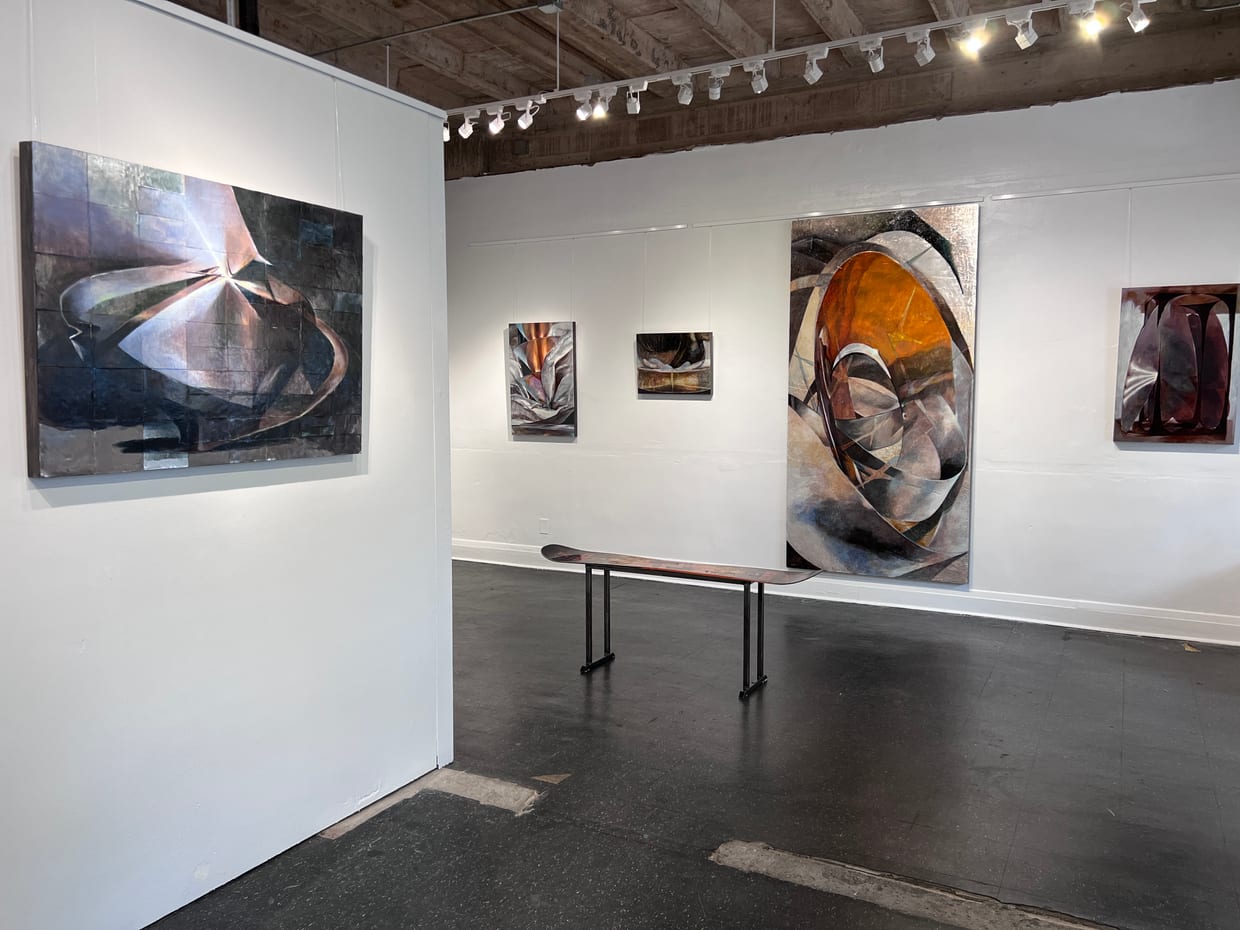 An art gallery interior with white walls displaying several large abstract paintings in dark metallic and orange tones, a wooden bench in the center, and exposed ceiling with track lighting.