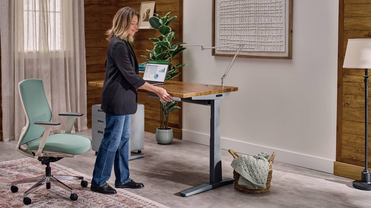 A woman raising an UPLIFT height-adjustable desk to standing height in a wood-paneled home office, a laptop on the desktop and an ergonomic task chair beside it.