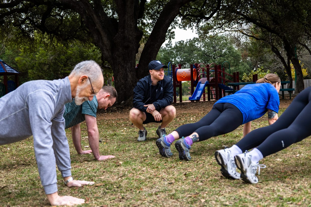 A fitness trainer crouches and coaches three clients doing push-ups on grass in a park with a playground in the background.
