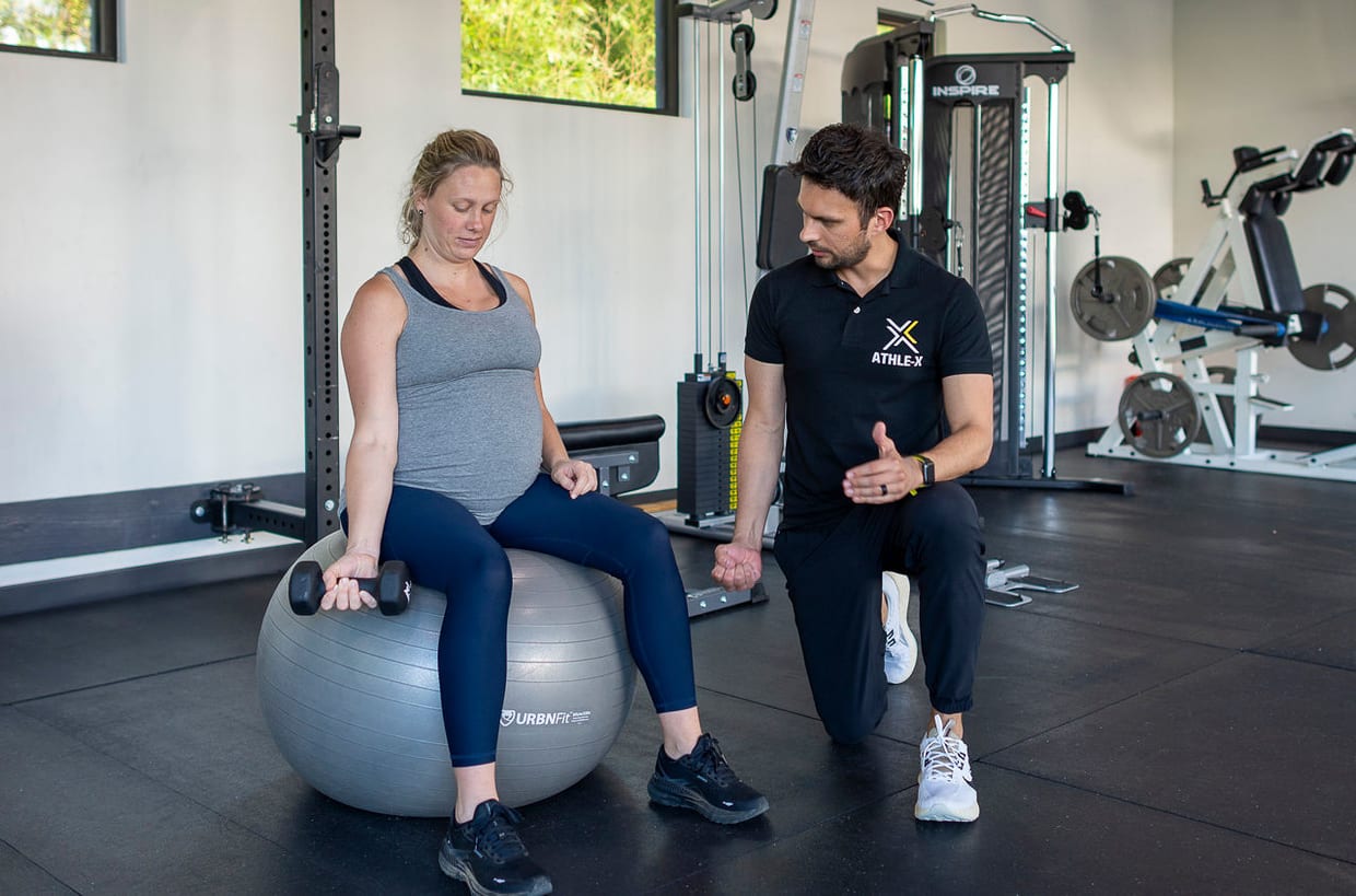 A personal trainer kneels beside a pregnant woman sitting on a stability ball holding dumbbells inside a well-equipped gym.