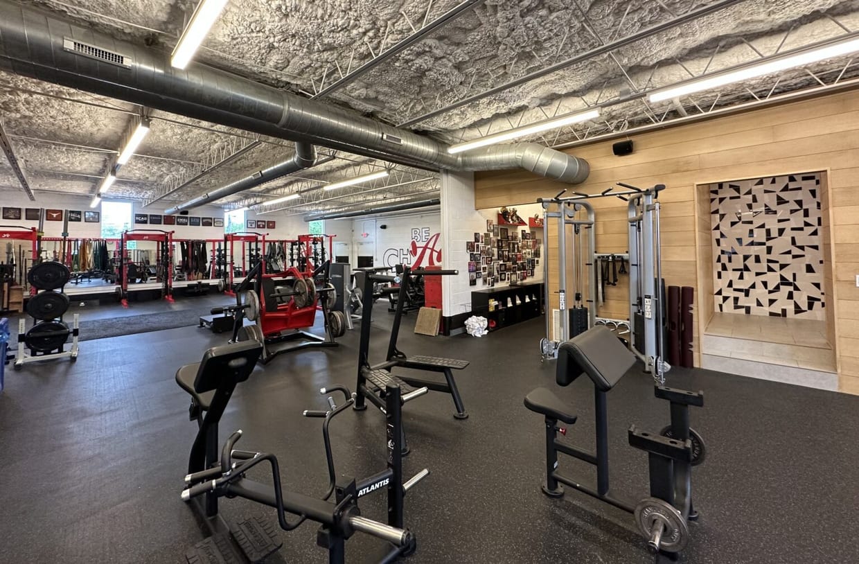 A spacious gym interior with black rubber flooring, rows of weight machines and red power racks, exposed ductwork ceiling, and a wood-paneled accent wall with framed photos.