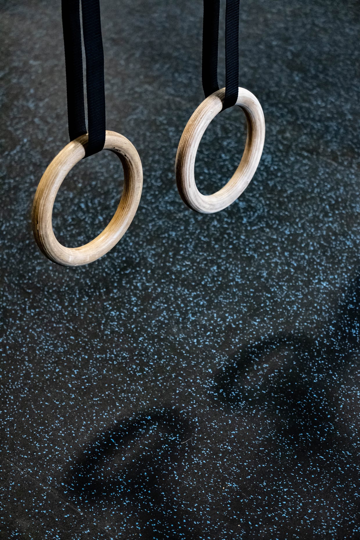 Two wooden gymnastic rings hang from black straps above a dark speckled rubber gym floor.