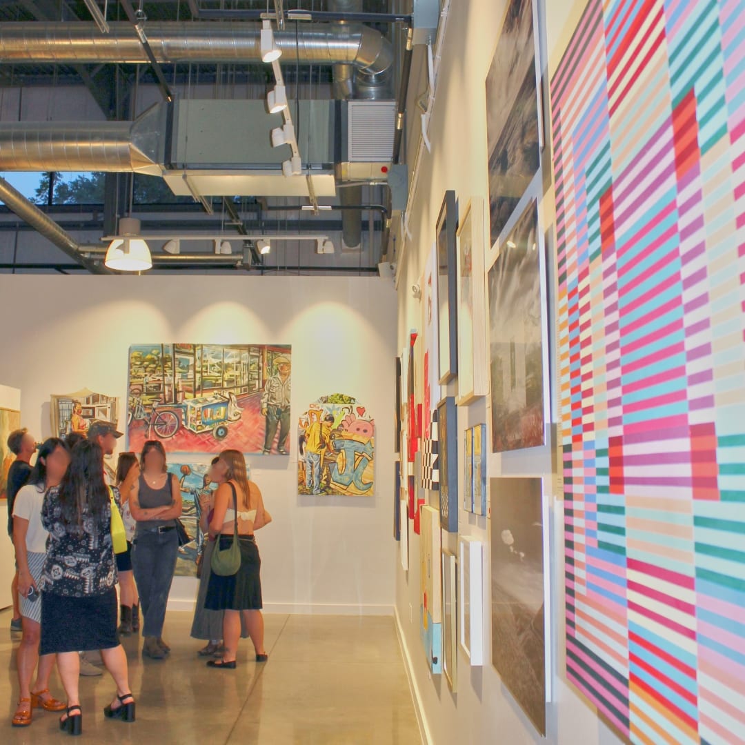 A group of visitors stands and converses inside a contemporary art gallery with multiple colorful paintings displayed on white walls under track lighting and exposed ductwork.