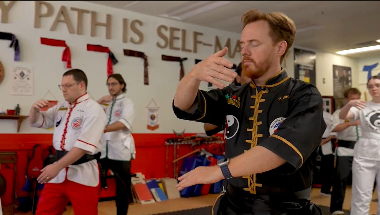 A martial arts instructor in a black and gold uniform leads a group of students through a technique drill inside a dojo with a motivational wall sign.