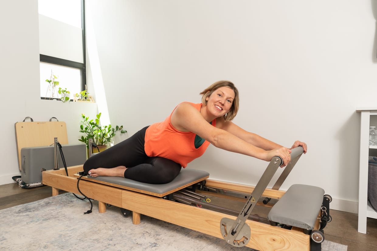 A smiling woman in an orange tank top and black leggings performs a stretch on a wooden Pilates reformer in a bright, plant-decorated studio.