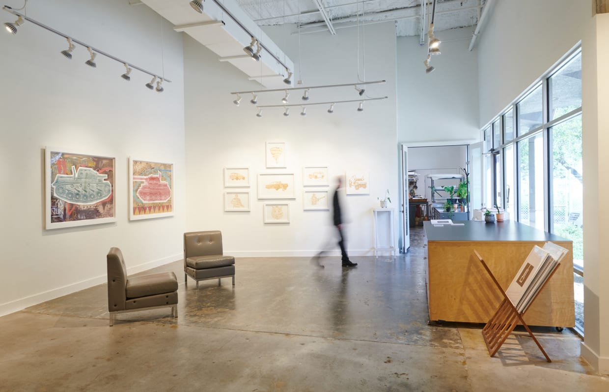 A bright contemporary art gallery interior with framed artwork on white walls, polished concrete floors, two chairs, a reception desk, and a motion-blurred person walking through the space.